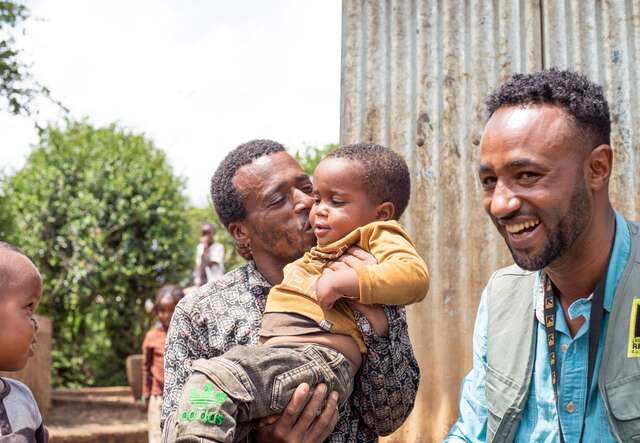 Eine Person, die ein Kind liebevoll im Arm hält, während eine andere Person daneben sitzt und lächelt.